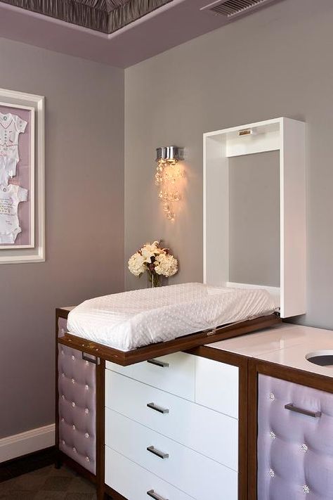 Glamorous gray and purple nursery features a concealed changing table that drops down over a dresser featuring white drawers accented with satin nickel pulls flanked by purple tufted upholstered cabinets located under diaper holes cut out from white countertops lit by a crystal beaded wall sconce mounted on gray walls. Lavender Nursery, Nursery Interior Design, Baby Changing Table, Dream Nurseries, Baby Changing, Everything Baby, Modern Nursery, Murphy Bed, Kids' Room