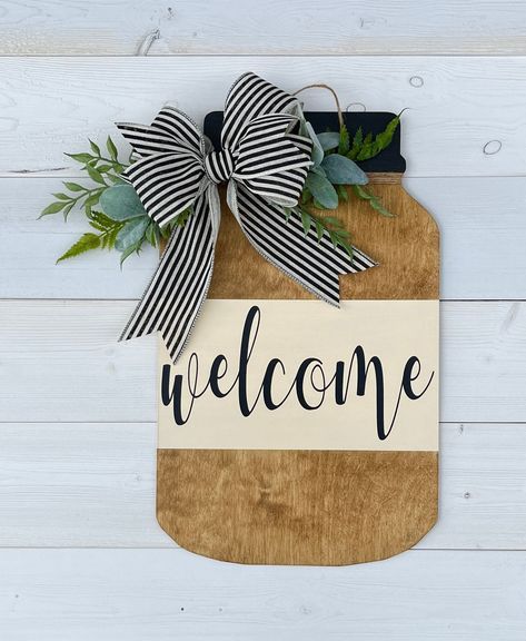 "A unique Year-Round Farmhouse style wooden mason jar that would bring a beautiful look to your front door. Stained with an \"Americana\" wood stain and painted with a milky white acrylic paint. The texts are done with a black premium permanent Vinyl. This lovely mason jar is also accented with a unique black/ivory stripe ribbon, and a touch of greenery to give it the ultimate farmhouse look. And to protect the wood, it is coated with an outdoor multi-surface water proofer. The measurements are Mason Jar Signs Wooden, Mason Jar Door Hanger Ideas, Ultimate Farmhouse, Mason Jar Door Hanger, Mason Jar Sign, Everyday Crafts, Front Door Farmhouse, Shop Door, Round Signs