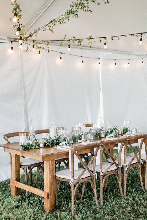 White wedding tent with wood harvest tables set with greenery and white pillar candles. Farm Table Garland Wedding, Harvest Table Greenery Wedding, Simple Harvest Table Decor Wedding, Greenery Farm Table Wedding, Harvest Table Wedding Centerpieces, Harvest Tables Wedding, Harvest Wedding Table, Harvest Table Decor Wedding, Outdoor Tent Wedding Reception Simple