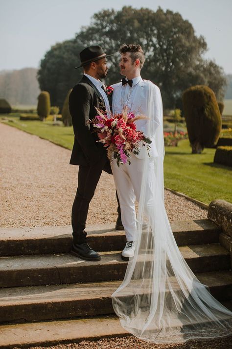 Groom in a black suit and fedora hat standing with his partner at Prestwold Hall Barns in a white groom suit and cape holding a vibrant red bouquet Wedding Suit With Veil, Unconventional Wedding Suit, Mens Wedding Suit With Cape, Wedding Suit With Cape, Celestial Wedding Suit, Fancy Wedding Suit, Enby Wedding, Gay Wedding Outfits, Groom In White Suit