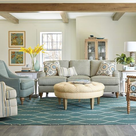 This charming living room has cream walls that seamlessly blend with the rustic charm of the natural wood beams above. The light gray sofa adorned with pillows that match the hues of the surrounding chairs sits on top of a large teal rug with white accents. The art features complimentary colors that help tie the space together. #KnoxvilleInteriorDesigner #InteriorDesign #HomeDecor #FamilyRoomDesign Natural Wood Beams, Classic Living Room Furniture, Light Gray Sofa, Light Gray Sofas, Cream Living Rooms, Cream Walls, Teal Rug, Classic Living Room, Sofa Upholstery