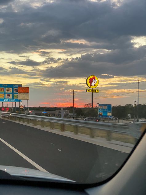 The picture is taken from the passenger seat of a car, looking out over a hot pink sunset far in the distance. In the foreground is a Highway sign listing local places to stop for food at the next exit (Taco Bell, McDonald’s, etc.) and a large sign for the gas station chain Buccee’s Bucees Texas Wallpaper, Texas Life Aesthetic, Bucees Texas Aesthetic, Buccees Aesthetic, Galveston Texas Aesthetic, Texas Astethic, Texas Summer Aesthetic, Atx Aesthetic, San Antonio Texas Aesthetic