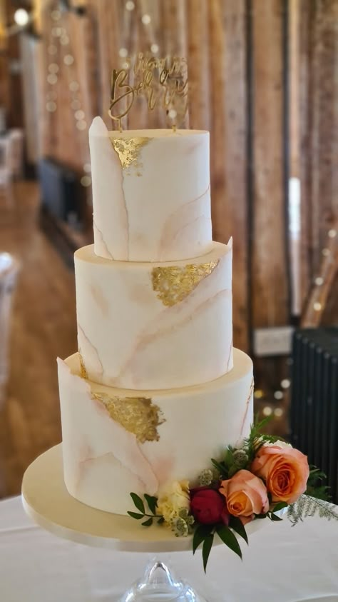 Three tier torn paper effect wedding cake with nude and blush dusting and edible gold leaf accents, sitting on clear glass cake stand with fresh flowers on the bottom tier in a barn setting with beams and fairy lights in the background Wedding Cake Champagne Color, Blush And Gold Wedding Cake, Honey Bee Wedding, Wedding Cake Gold Leaf, Bee Wedding Cake, Torn Paper Effect, Wedding Cake Marble, Cakes With Flowers, Orange Wedding Cake