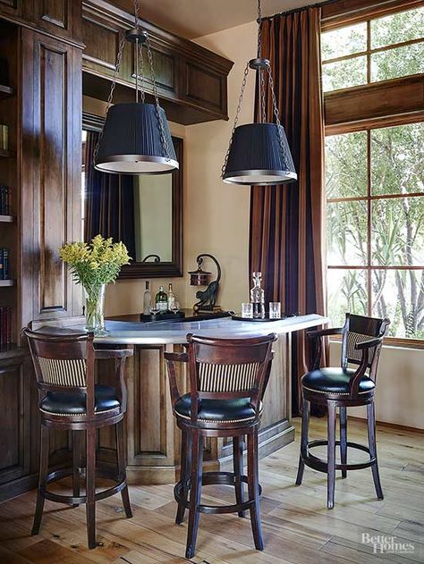 Extending out from a windowed corner, this handsome home bar takes its cue from the room's British-study leanings and paneled woodwork. A back-bar mirror, strikingly shaded pendant lights, and a white marble bar top draw attention to the drink station, which is outfitted with cozy leather seating. Bar With Window In Middle, Home Bar Window, Bar Pass Through, Corner Bars For Home, Bar With Window, Bar In Bay Window, Home Bar With Seating, Corner Wet Bar, Home Bar Antique Mirror