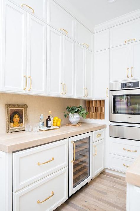 Elegant white and gold kitchen pantry boasts white shaker cabinets accented with brass pulls and holding a glass front wine fridge over a rustic wood floor and beneath a beige countertop matching a gold raffia backsplash. Rustic Wood Floor, White And Gold Kitchen, White Kitchen Pantry, Small Kitchen Pantry, White Pantry, Rustic Wood Floors, Pantry Cabinets, White Shaker Cabinets, Wood Floors Wide Plank