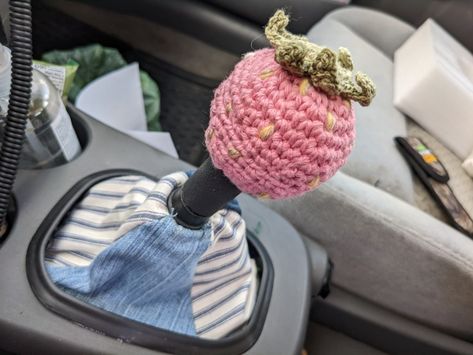 A gear shift is shown in a gray car, the cloth boot cover is alternating blue jeans fabric and Blue and white striped fabric. The round gear knob has a crocheted cover, it is shaped as strawberry and is light pink with yellow seeds and green leaves on top. Car Shift Cover, Diy Crochet Car Accessories, Chevy Car Accessories, Crochet Stick Shift Cover, Crochet Car Shifter Cover, Crochet Car Gear Shift Cover, Cute Car Themes, Skullcandy Crusher Evo Aesthetic, Crochet Car Cover