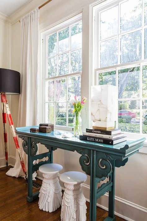 Chic contemporary living room features two white fringe stools placed beneath a peacock blue chinoiserie console table placed in front of a window covered with white curtains. Console Table Desk Behind Couch, Console Table Window, Console In Front Of Window, Table In Front Of Window Living Room, Console Table In Front Of Window, Window Console Table, Table Under Window, Table In Front Of Window, Executive Office Design Interior