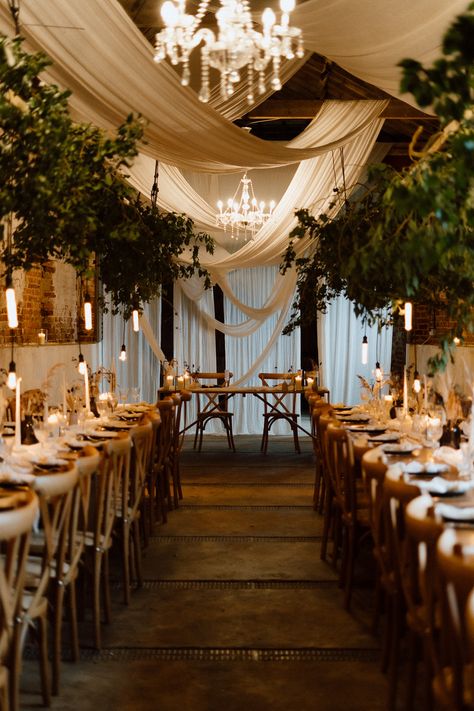 This is showing a barn wedding dining area. The overall vibes are rustic luxury. On the ceiling are glass chandeliers, white drapes hanging across the ceiling, directly above the wooden long dining tables are hanging boards with ivy on them. On the tables are white table runners, vases, candlesticks, tableware and glassware. In the middle of the two rows of dining tables is a sweet heart table at the end. Wooden Crossback Chairs Wedding, Low Ceiling Draping Wedding, Wedding Ceiling Drapery, Wedding Decor Hanging From Ceiling, Wedding Ceiling Decorations Draping, Green Draping Wedding, Curtain Ceiling Design, Wedding Drapes Ceiling, Black Draping Wedding