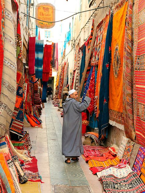 a street open market in essaouira,morocco with colourful carpets and a sale person • Buy this artwork on home decor, stationery, bags et more. Moroccan Street, Morocco Essaouira, Design Marocain, Moroccan Culture, Morocco Travel, Moroccan Decor, Carpet Colors, Moroccan Style, Places Around The World