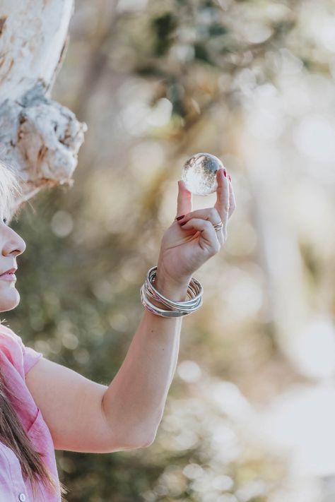 The image shows a close-up of a woman's hand holding a small, clear crystal ball between her fingers. She is wearing multiple silver bangles on her wrist, and the background is blurred, showing hints of greenery and soft light. The scene gives a sense of focus and contemplation, with the crystal ball catching the light and reflecting the surroundings. Health Coach Branding Photoshoot, Wellness Coach Branding Photoshoot, Holistic Health Coach Photoshoot, Therapist Branding Photos Outdoor, Holistic Health Coach Branding, Wellness Photography, Personal Brand Photoshoot, Learn Business, Personal Branding Photography