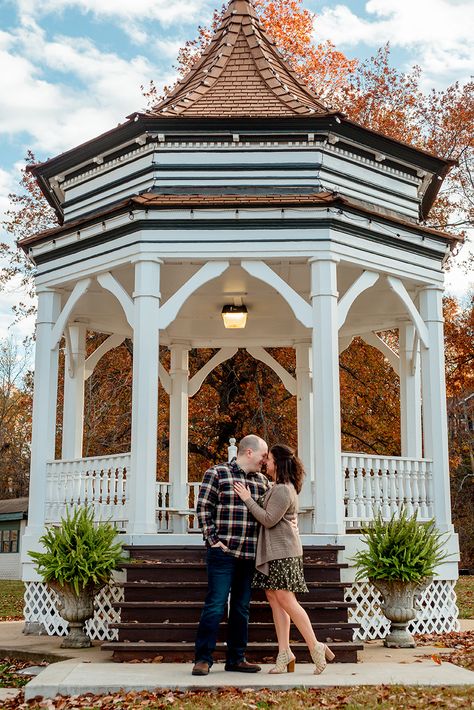 A heterosexual couple stands under a gazebo, foreheads pressed together and looking into each other's eyes. The sky behind them is blue with a few clouds, and the leaves are bright orange. Gazebo Couple Photoshoot, Gazebo Couple Pictures, Engagement Photos Gazebo, Gazebo Engagement Pictures, Small Town Couple Photoshoot, Gazebo Photoshoot Ideas, Small Town Engagement Photos, Small Town Photoshoot, Town Photoshoot