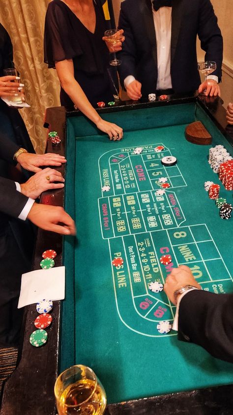 A craps table surrounded by tux covered hands, wine glasses, ladies in evening gowns. Wine glass in the lower corner, dice and chips on the table as play is in session Craps Table, Imaginative Writing, Casino Room, Surprise 60th, Casino Table Games, Green Room Decor, Casino Party Games, Casino Birthday, Casino Table