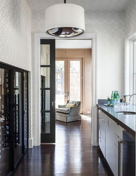 A black pocket door with glass panels opens to a long butler's pantry featuring walls clad in white and gray trellis wallpaper fitted with a built-in wine cellar finished with clear glass sliding doors facing white cabinets fitted with a pair of stainless steel mini refrigerators topped with gray quartz countertop framing a round sink and gooseneck faucet placed under windows illuminated by a white drum pendant. Stainless Steel Door Design, Grey Trellis Wallpaper, Neutral Family Room, Glass Pocket Door, Sliding Pantry Doors, Glass Pocket Doors, Steel Door Design, Stainless Steel Door, Glass Panel Door