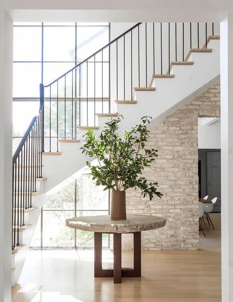 Minimalist foyer boasts a round concrete table topped with a potted plant and positioned in front of white staircase finished with an iron handrail and spindles and wood treads illuminated by natural light from a large black factory window accented with wall half covered in brick. Boho Floral Nursery, Staircase Window, Foyer Staircase, Floral Nursery Decor, Mediterranean Style Home, Interior Finishes, Mediterranean Home Decor, Take The Stairs, Mediterranean Decor