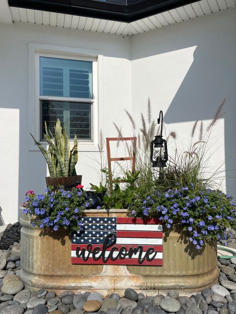 A variety of zone, sunlight, and soil plants inside a livestock tank with added visual interest such as a rustic bucket, a ceramic hen nestled in the plants, a small shepherd’s hook with an antique lantern, and a patriotic sign hanging on the front. At night, the display, which sits between the driveway and front door, is lit up by a solar spotlight. Repurposed Water Trough, Water Trough Ideas Backyards, Stock Tank Repurposed, Cow Trough Garden, Trough Flower Planters, Trough Flower Garden, Water Trough Flower Planter, Water Tank Planter Ideas, Metal Horse Trough Planters