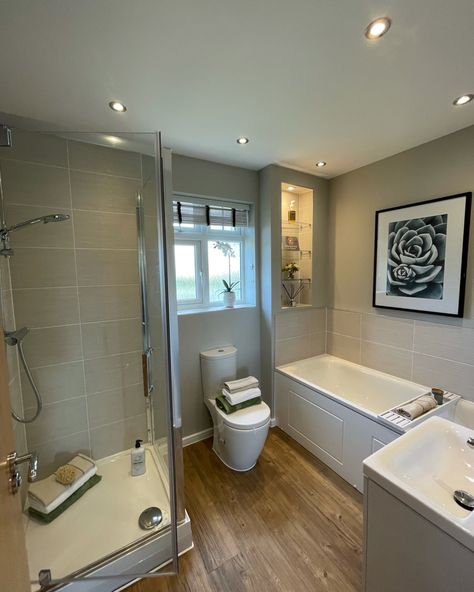 A show home bathroom, that is made up for a walk in shower, a separate bath, a toilet and a wall hung sink. The colour scheme is cream and brown, with a wooden floor and cream tiles. The bathroom suite is white. There are shelves built into the wall which are decorated with a plant Bathroom With Cream Tub And Toilet, Countryside Bathroom, Grey And Cream Bathroom, Beige And Grey Tiles Bathroom, Cream Toilet And Sink, Cream Bathroom Ideas, Bathroom With Almond Tub And Toilet, Bathroom Tile Brown And Cream, Cream Bathroom