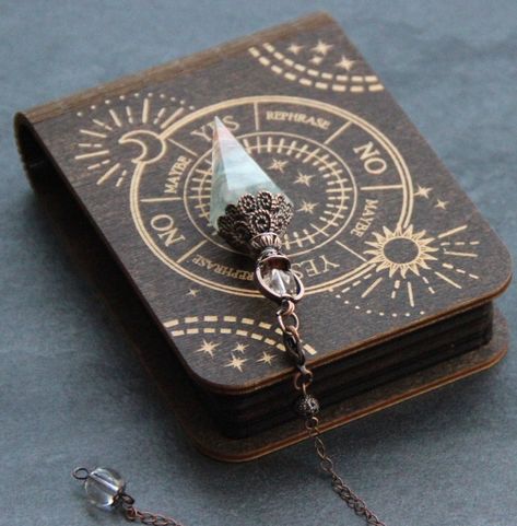 "Our divination set includes everything you need to get started with pendulum divination: a Pendulum and a wooden box with a Pendulum board lid which doubles as a pendulum holder, keeping your crystal pendulum safe and secure when not in use. You have the option to select between three pendulums to complete your set or to buy only the box as a home to your favorite pendulum. This dowsing kit is also great gift for those who are just starting out with divination and want to explore this ancient a Pendulum Holder, Magician Wand, Pendulum Divination, Diy Kandi Bracelets, Pendulum Board, Crystal Pendulum, Inner Wisdom, Paper Gift Box, Green Witch