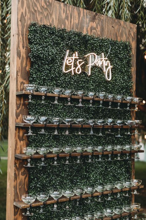 A dark stained walnut champagne wall with wide mouthed champagne glasses, behind it is foliage, at the top there is a LED sign that says "lets party" Wine Wall Display Party, Champagne And Beer Wall Wedding, Beer Tap Wall Wedding, Welcome Champagne Wall, Champagne Flute Wall, Rustic Champagne Wall, Welcome Shots Ideas, Shot Wall Wedding, Wedding Beer Tap