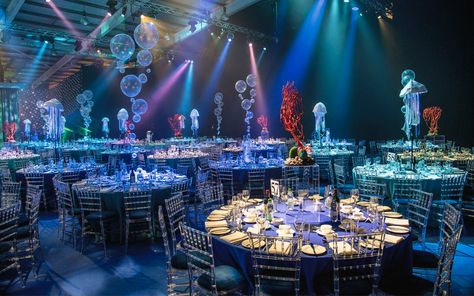 To access the dining area guests made their way through an opulent deep wall of fabric columns, emulating waves and shafts of light in the sea. Dining tables were dressed in four different shades of blue cloths, surrounded with clear perspex chairs. Bubble, coral and internally illuminated jelly fish table centres combined with creative under the sea lighting brought the space to life and left guests utterly in awe as 3mtr jellyfish acrobats danced the night away over the top of the tables. Under The Sea Gala, Explore Images, Sea Wedding Theme, Grad Dinner, Debut Theme, Under The Sea Decorations, Gala Night, Prom Themes, Sea Cakes