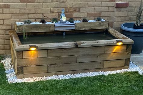 A wooden raised pond with a wood shelf and metal waterfall feature. Alpine plants and a Buddha statue are situated on the shelf above the pond. Raised Wooden Pond, Raised Pond With Waterfall, Raised Ponds, Homemade Waterfall, Above Ground Pond, Raised Pond, Pond Kits, Building A Pond, Diy Pond