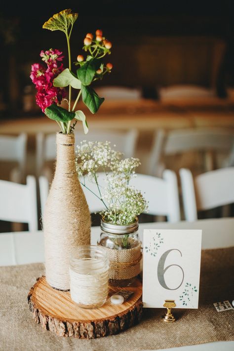 Rustic Center pieces, twine wrapped wine bottles, mason jars, paper cards, wood rounds  Rustic Grace Estate, Van Alstyne, TX  Photocred: Two Pair Photography Wedding Centerpiece With Wine Bottles, Wine Bottle Mason Jar Centerpiece, Rustic Wine Bottle Centerpieces, Boho Wine Bottle Centerpieces, Wine Bottle Centrepiece, Whiskey Bottle Wedding Centerpieces, Wine Bottle Diy Wedding, 21st Birthday Centerpieces, Lilac Wedding Themes