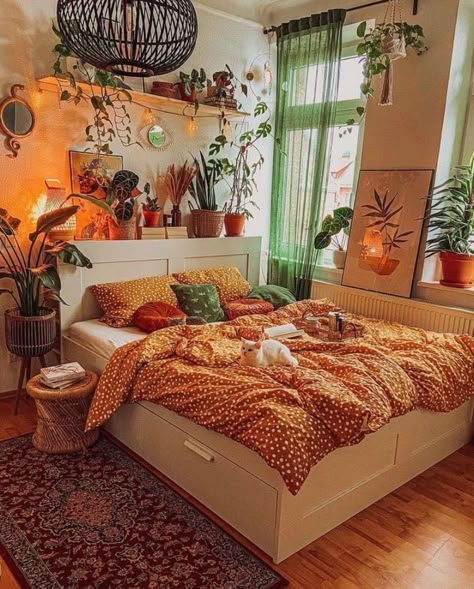 Bedroom with orange comforter, warm, pink Himalayan salt lamp lighting, with green accents from many plants and sheer green curtain over the window. Warm  brown wooden floor and white wood bed frame. Unique Bed Spreads, Jungalow Bedroom Bohemian, 70s Pastel Decor, Earth Toned House Decor, Colorful Bedroom With Plants, Bed Rooms Ideas Maximalist, Wooden Wall Bedroom Ideas, Whimsy Bedroom Decor, Mix Match Side Tables Bedroom