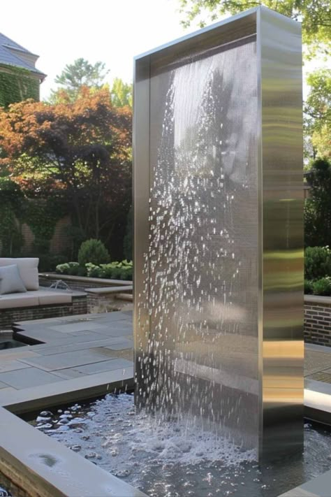 A sleek, modern metal fountain with water cascading down a vertical panel, set in a contemporary patio with outdoor seating and lush greenery. Modern Fountain Design, Swimming Pools Backyard Landscape, Unique Backyard, Backyard Features, Villa Landscape, Modern Water Feature, Modern Fountain, Outdoor Water Features, Contemporary Garden Design