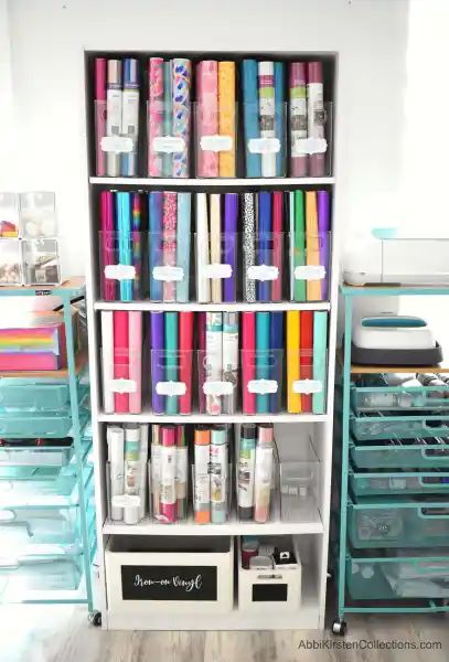 A white, tall bookshelf holds Cricut vinyl in clear storage bins organized by type. Labels make finding what you need easy and hassle-free. Vinyl Roll Storage Ideas Diy, Craft Room Printables, Storage For Craft Room, Cricut Office, Craft Storage Ideas For Small Spaces, Office Craft Room Combo, Cricut Storage, Organizing Office, Craft Storage Solutions