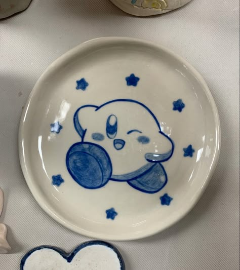 small to medium sized white porcelain plate , in the middle there is a handpainted Kirby. A character from nintendo, the kirby is encircled by 8 small stars Kirby Ceramic Mug, Kpop Pottery Ideas, Kuromi Pottery Painting, Cute Pottery Aesthetic, Porcelain Mug Painting, Cute Ceramic Bowl Designs, Kirby Clay Tray, Pottery Designs Plate, Anime Pottery Ideas