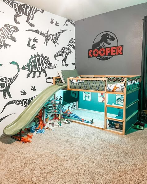 Surprise your son with a fun Jurassic World-themed bedroom. The adorable tiny bunk bed with olive sheets features a slide attached to its side. Complete the space with two dinosaur-themed accent walls. Dinosaur Room Ideas Toddler, Kids Dinosaur Bed, Small Dinosaur Bedroom Ideas, Dino Inspired Bedroom, Toddler Boy Room Decor Dinosaur, Dinosaur Boy Room Ideas, Jurassic World Room Decor, Dinosaur Bedroom Ideas For Boys, Little Boys Dinosaur Bedroom