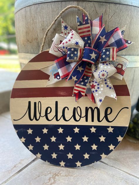 "Add a little patriotic farmhouse flair to your front door with this distressed Fourth of July or Memorial Day door hanger. Each door hanger is made from Baltic Birch wood and measures 16\" in diameter and .25\" deep and comes with a jute rope hanger. Back of sign is finished with stain or paint. Wording and designs are made from outdoor vinyl.  Greenery and bows are attached using staples.  Both front and back of signs are sealed for outdoor use, but it is recommended for indoor use or displayed on door under a covered porch or patio.   Each sign will vary slightly due to the way the wood will accept the paint and stain.  This is what will give each sign its unique and custom look.   Each sign is made at the time order is received.  Multiple quantities are listed so more than one person c Red White Blue Door Hanger, 4th Of July Door Hanger Diy, Patriotic Diy Crafts, Diy Patriotic Decor, Patriotic Door Decorations, Fourth Of July Door Hangers, 4th Of July Door Hanger, 4th Of July Crafts, Fourth Of July Wreath