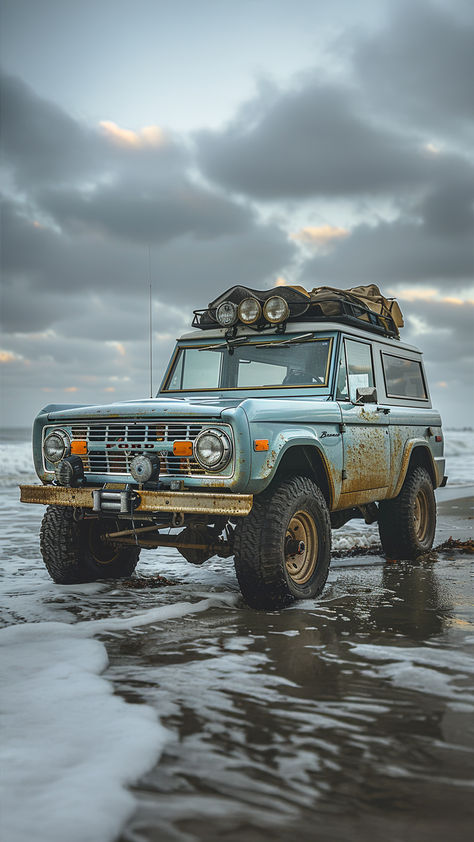 Vintage blue Ford Bronco parked on a beach with waves touching the tires. The SUV is equipped with roof-mounted lights and off-road tires, set against a cloudy sky, embodying a sense of adventure and readiness for beach camping. Blue Bronco, Old Ford Bronco, Adventure 4x4, Broncos Pictures, Chevy K10, American Pickup Trucks, Classic Bronco, Early Bronco, Classic Ford Broncos