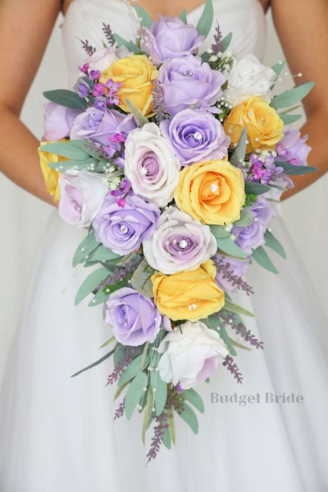 This lovely assortment of lavender, wisteria and yellow roses is bound to turn heads on your wedding day. This lovely cascading brides’ bouquet from the is an absolutely breathtaking arrangement for any bride to be, in any season. Bright greenery and lavender and lilac accents with pearls complete the look and add a delicate realism to this beautiful bouquet. This bouquet is 10” wide, 18” long, and is crafted by our in-house florists without using a foam base to anchor the flowers to the bouquet Yellow Roses And Lavender Bouquet, Garden Theme Bouquet, Purple And Yellow Bridal Bouquet, Purple And Yellow Wedding Ideas, Wisteria Flower Bouquet, Lavender And Yellow Wedding Theme, Lemon And Lavender Wedding, Rapunzel Flower Bouquet, Purple And Yellow Wedding Flowers