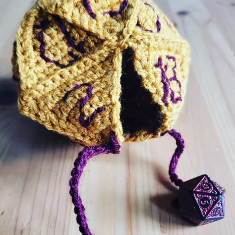 A crocheted bag shaped as a 20 sided dice in gold and dark purple with purple ties to close. A small purple and black 20 sided dice sits next to the bag. Crochet D20 Pattern, Dice Crochet, D20 Crochet Free Pattern, Crochet Fuzzy Dice, D20 Crochet, Crochet Dnd Dice Bag, Crochet D20, Crochet Dice, D&d Crochet