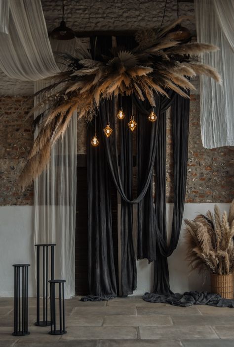 This image shows a barn wedding ceremony area. The main focus is on a hanging dried floral installation. The installation consists of normal cream pampas leaves and dark black ones, creating a depth in the installation. There are also uniquely shaped light bulbs hanging from the installation. Behind the installation are black and white drapes hanging across the venue ceiling and down the wall. Below the flowers are black plinths. Black Wedding Drapery, Black Wedding Draping, Suspended Floral Arrangements, Fabric Hanging From Ceiling, Yalda Decor, Backdrop Ideas For Wedding, Diy Ceremony Backdrop, Unique Backdrop Ideas, Chelsea Fabric