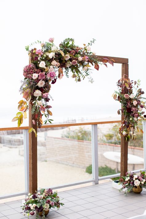 A rectangular, dark wood arch with asymmetrical purple, plum, and white floral decor.  #malibuwestbeachclub #malibuwestbeachclubwedding #delmarevents #malibuwedding #asymmetricalfloraarch #woodceremonyarch #ido #malibuwedding #malibubeachwedding #plumarchflorals Mauve Wedding Arch Flowers, Asymmetrical Floral Arch Wedding, Wood Arch With Flowers, Mauve Wedding Arch, Rectangular Arch Wedding, Asymmetrical Floral Arch, Wood Arches Wedding, Rectangular Wedding Arch, Dark Purple Bridesmaid Dresses