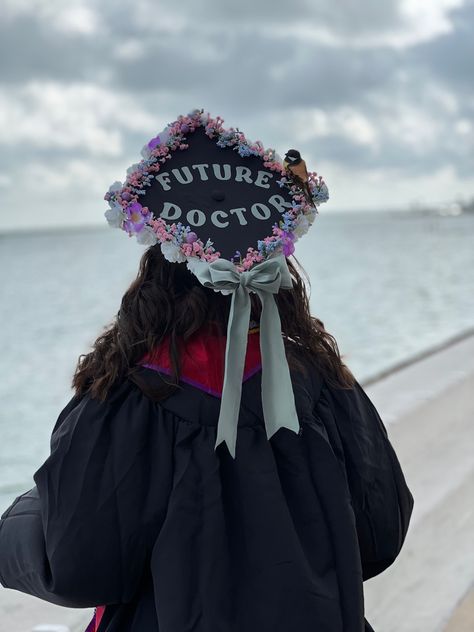 Picture of graduation cap with white roses on the bottom and pink, purple, and blue flowers on the top lining the cap, and surrounding the words “Future Doctor” in mint green in the center. A fake bird perches on the edge of the caps flowers. A large mint green bow in the same fabric and color shade as the words above is on the edge of the cap pointing towards her graduation gown Graduation Cap Designs Medical School, Future Doctor Grad Cap, Future Doctor Graduation Cap, Graduation Cap Designs College Medical, Pre Med Graduation Cap, Medical Graduation Cap, Biology Graduation Cap, Graduation Cap Diy, Medicine Graduation