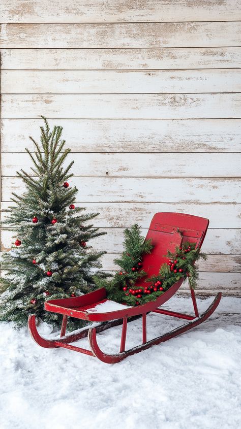 A red vintage sled adorned with greenery and holly berries on a wooden porch, beside a small Christmas tree, in a snowy Wooden Backdrop, Rustic Front Door, Luxury Christmas Decor, Front Door Styles, Little Christmas Tree, Vintage Sled, Wooden Backdrops, Rustic Porch, Cozy Rugs