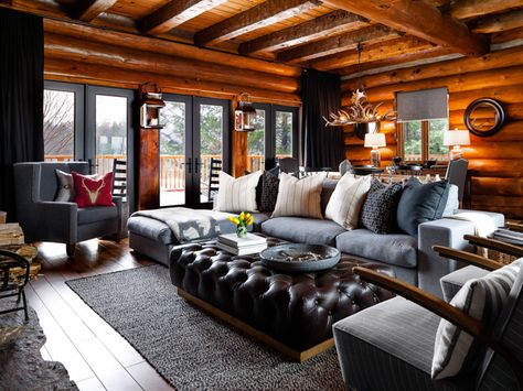 Dressed with an oversized Casa Life sofa and a leather ottoman, the great room scene is anchored by a large gray rug that seems to float, like an island, in front of the stone-built fireplace. Our luxe lodge vibe mixes wood finishes, leather detail, and gray linen to elicit a soft and relaxing vibe. Black accents, via drapery and accessories, anchors the scheme. RELATED: 22 Living Room Before and Afters Log Home Living Room, Wood Paneled Walls, Log Cabin Interior Design, Paneled Walls, Log Cabin Furniture, Cabin Interior Design, Log Cabin Living, Log Home Living, Log Cabin Interior