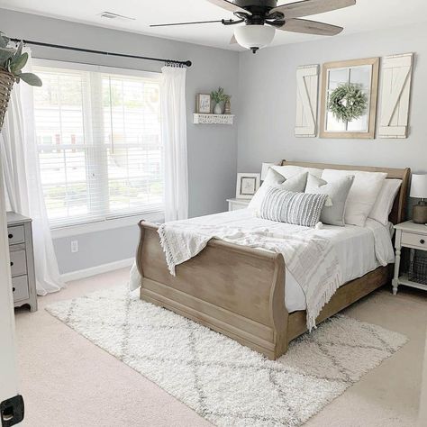 White window shutters and a wood-framed mirror hang on a light grey bedroom wall above a wooden sleigh bed. A light grey textured bolster leans against grey pillows on the white coverlet. A white rug with grey stripes is laid on the off-white carpet. Carpet White Walls, White Walls Bedroom, Light Grey Carpet, Wooden Sleigh Bed, Cozy Grey Bedroom, Bedding Farmhouse, Light Gray Bedroom, Gray Bedroom Walls, White Coverlet