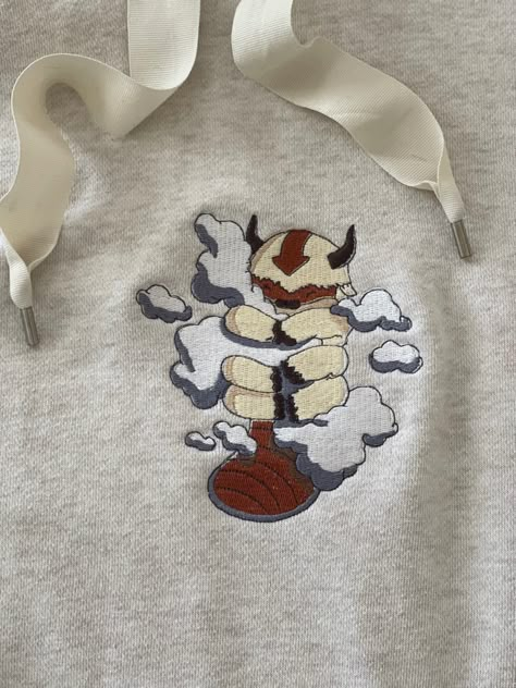 A cream colored sweatshirt with a machine embroidered sky bison named appa. from avatars the last Airbender he is hugging clouds. Avatar The Last Airbender Hoodie, Avatar The Last Airbender Sweatshirt, Appa Embroidery Pattern, Appa Sweater, Embroidery Designs On Hoodies, Machine Embroidered Clothes, Avatar The Last Airbender Embroidery, Embroidery Avatar, Appa Hoodie