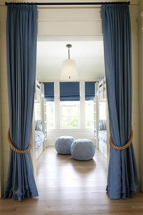 Blue curtains with rope tie-backs open to a bunk room filled with two blue tufted poufs flanked by white bunk beds dressed in blue bedding placed in front of a row of windows covered in denim blue roman shades. Curtain Color Ideas, Denim Curtains, White Bunk Beds, Rope Tie, How To Dress A Bed, Blue Curtains, Bunk Room, Blue Home Decor, Home Curtains