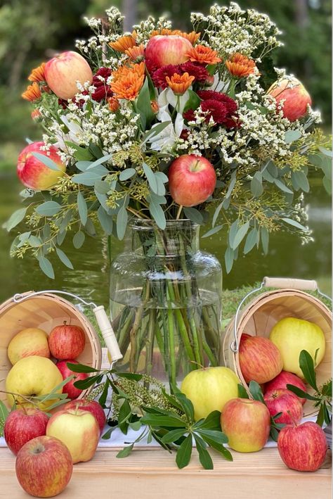 A floral arrangement adorned with mums, eucalyptus, and apples along with bushel baskets overflowing with apples create the perfect centerpiece for an Apple Harvest Tablescape. Pumpkins And Apples Decor, Apple Display Table, Fall Apple Party Decorations, Apple Harvest Party Ideas, Apple Boquet, Red Apple Centerpiece Ideas, Fall Apple Birthday Party, Apple Theme Centerpieces, Apple Festival Decorations