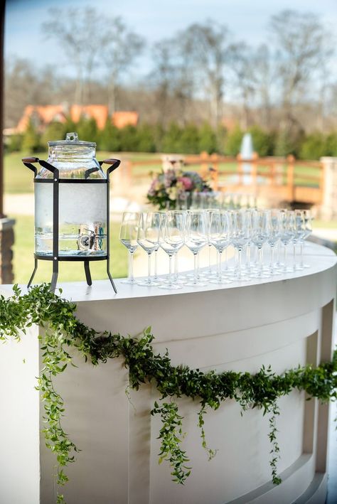 A white bar with swag of greenery sits in front of a green space with fountain and bridge. The cocktail bar area is set up with glassware and a beverage dispenser. Wedding Alcohol Bar, Wedding Cocktail Hour Ideas, Bar Area Decor, Cocktail Hour Ideas, Outdoor Wedding Bar, Outdoor Cocktail Party, Cocktail Hour Decor, Wedding Alcohol, Wedding Cocktail Hour