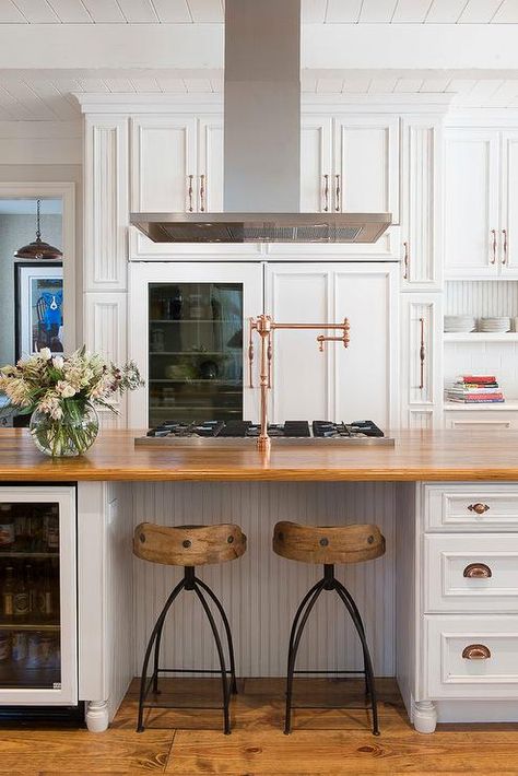 Stunning kitchen features white raised panel cabinets adorned with copper hardware framing a glass door refrigerator. Farmhouse Kitchen Pot Filler, Island Pot Filler, Stove In Island With Hood, Kitchen Island Hood Vent, Stove In Kitchen Island, Island Hood Vent Ideas, Kitchen Island Stove, Kitchen Island Hood, Stove In Island