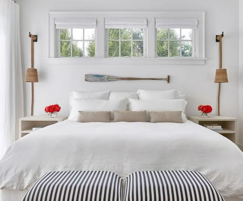 Cottage bedroom features three windows dressed in white roman shades placed over a decorative oar lining a wall above a bed dressed in white bedding and taupe pillows flanked by white linen nailhead open nightstands illuminated by rope wall sconces. Remote Shades, Windows Apartment, Window Above Bed, Modern Coastal Bedroom, Lake House Bedroom, Beach House Tour, Beach Themed Bedroom, Beach House Bedroom, Beach Bedroom Decor