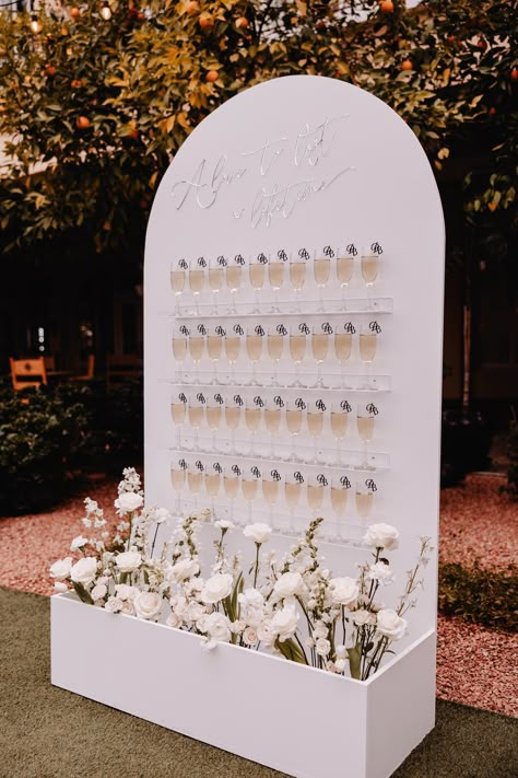 There's no better way to get the party started with your guests as they enter your cocktail hour or reception than with an interactive wedding champagne wall! This classic white champagne wall with a custom quote at the top for the couple was the coolest touch to their big day! View more from this luxury Phoenix wedding captured by Jenaya Michelle Photography, an Arizona wedding photographer! Sip And Be Seated Champagne Wall, Champagne Seating Chart Wall, White Wedding Cocktail Hour, Cocktail Hour Entertainment Ideas, White Champagne Wall, Champagne Wall Diy, Shot Wall Wedding, Drink Wall Wedding, Champagne Bar Wedding