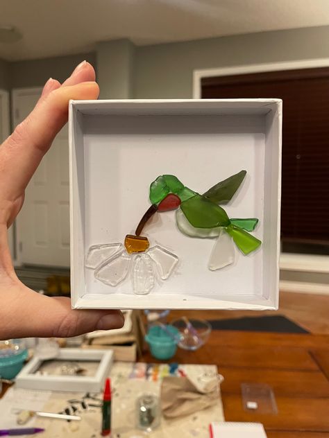 Hand holding up small shadow box. Inside shadow box are pieces of sea glass made in the shape of a daisy and a ruby throated hummingbird. Beach Glass Ideas, Sea Glass Hummingbird, Sea Glass On Canvas, Sea Glass Turtle Art, Sea Glass Canvas Art, Brown Sea Glass Art, Sea Glass Resin Art, Beach Glass Art Ideas, Sea Glass Animals