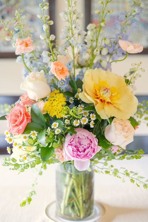 A pastel spring floral arrangement in a glass vase featuring soft pink peonies, yellow poppies, peach roses, delicate chamomile, and lush greenery. Perfect for weddings and spring events, this arrangement creates a romantic and whimsical vibe. Yellow Pink Blue Flower Arrangement, Pastel Rainbow Flower Arrangements, Pink Yellow White Flower Arrangement, Colorful Floral Arrangement, Blue Flower Arrangements, Shower Flowers, White Floral Arrangements, Green Centerpieces, White Flower Arrangements