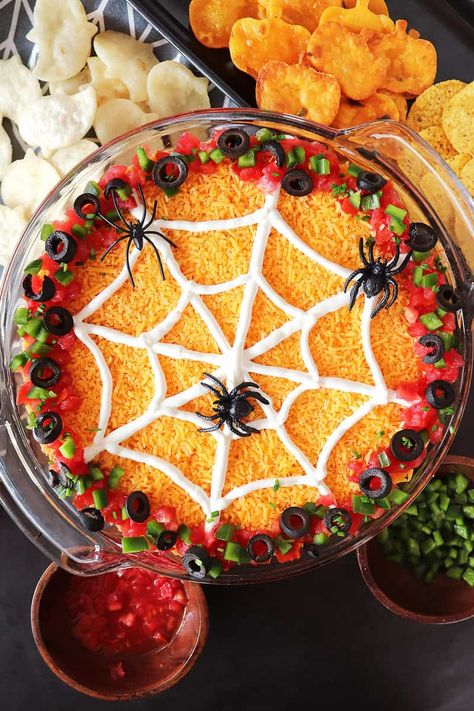 This Halloween 7-layer dip recipe is made with layers of refried beans, guacamole, salsa, taco-seasoned sour cream, and shredded cheddar cheese, topped with sliced black olives, diced green peppers, and tomatoes. A spooky sour cream spider web decorates the top and don't forget the spiders! Spider Web 7 Layer Bean Dip, Spider Web Dip Sour Cream, Halloween Bean Dip Spider Webs, Halloween Spider Dip, Spider Web Seven Layer Dip, Spider 7 Layer Dip, Spider Seven Layer Dip, Spiderweb Bean Dip, Spider Web Bean Dip